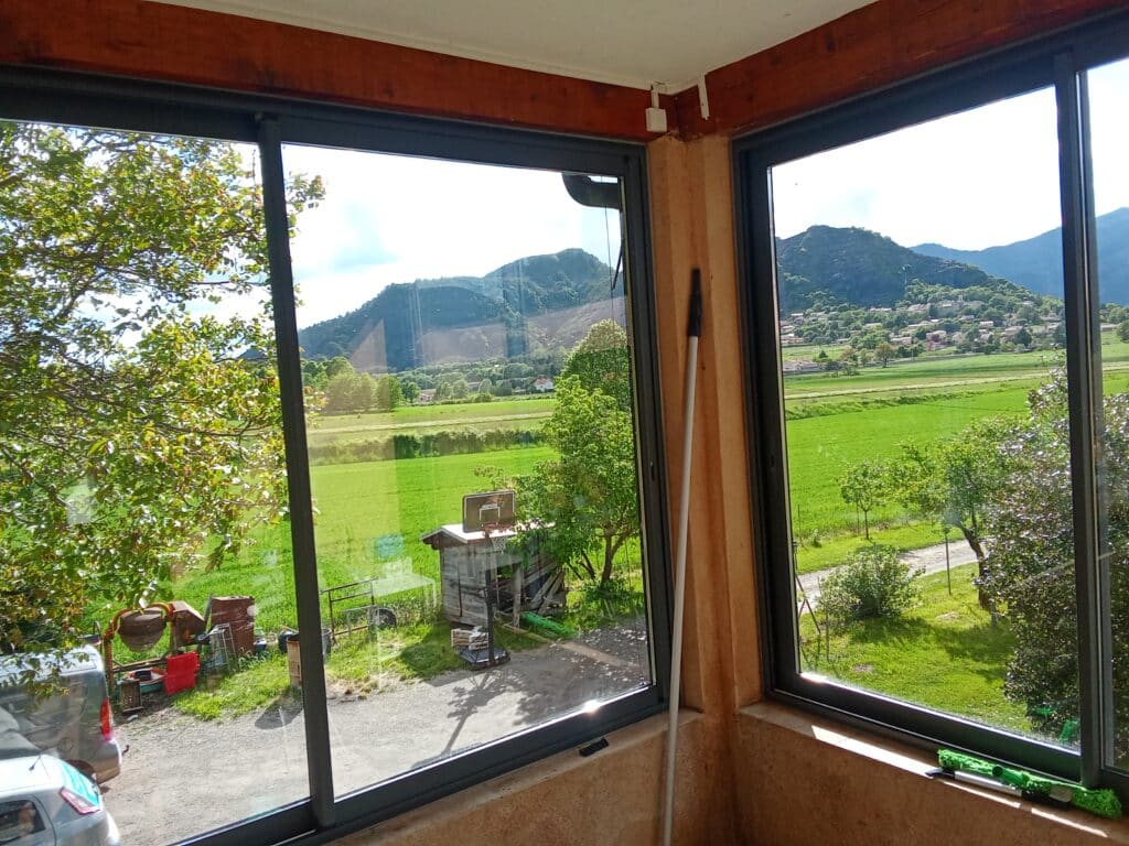 Vue Panoramique de Montagnes et Vallée via une Baie Vitrée Vue d'une vallée verdoyante et de montagnes à travers de grandes baies vitrées, avec un jardin au premier plan.